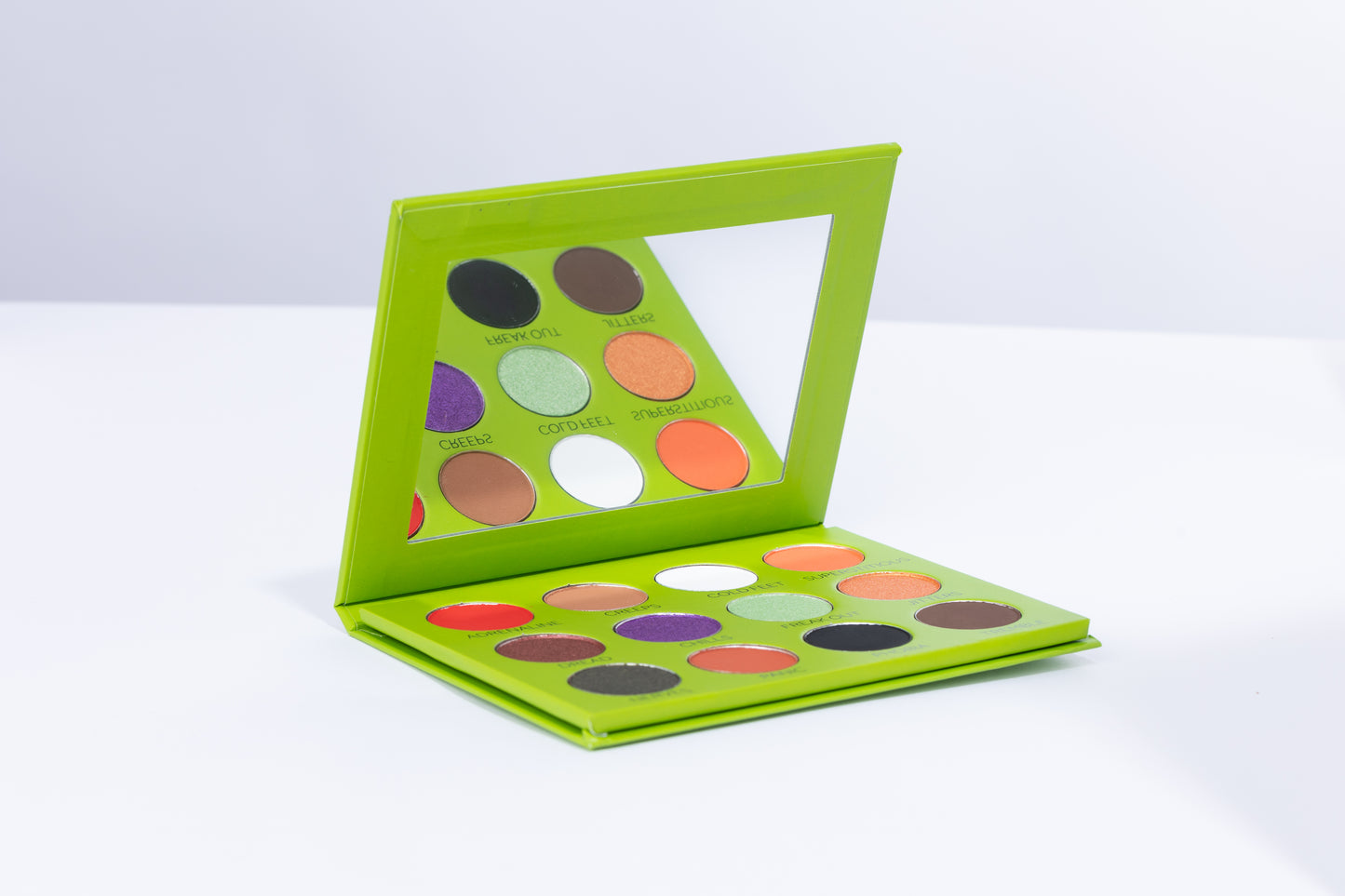 A Halloween-themed eyeshadow palette with a mirror, displayed in a green packaging, showcasing vibrant makeup colors.